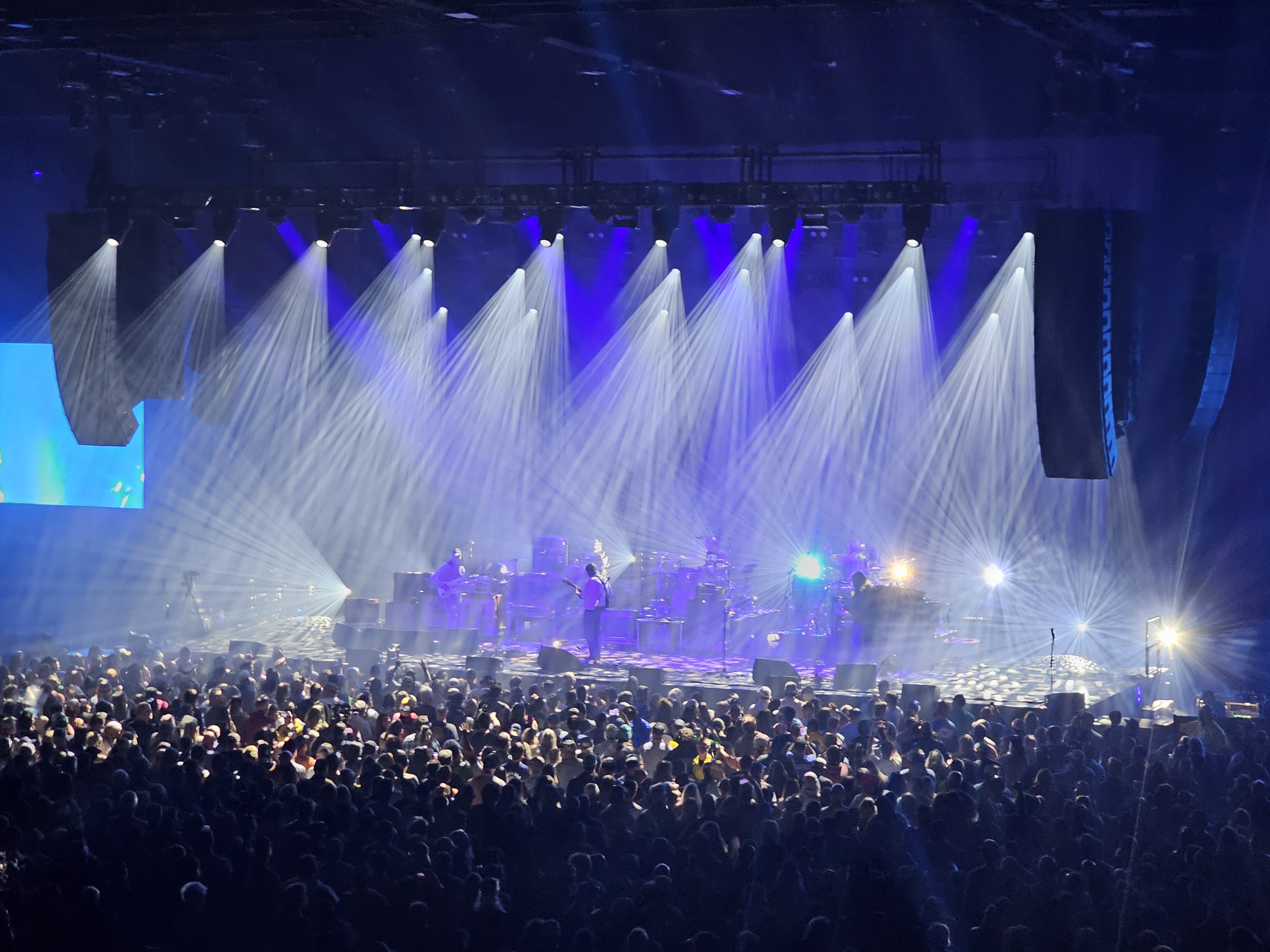 A large Widespread Panic concert at the Hard Rock Live Etess Arena in Atlantic City, New Jersey with a packed audience standing in front of a brightly lit stage. White and blue beams of light radiate dramatically from above, illuminating the band performing at center stage. The lighting creates a dynamic, atmospheric effect, while the crowd is silhouetted in the foreground. Large speaker stacks hang on both sides of the stage, and visual effects are projected on screens in the background.
